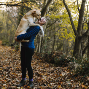 young blonde woman holds yellow lab dog while on a hike in the woods